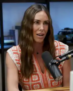 A portrait photograph of a woman with medium-length wavy brown hair wearing an orange and white shirt. She is sitting at a wooden table in front of a microphone.