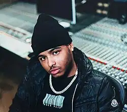 Jay Whiss wearing a black print t-shirt under a puffy black jacket, sitting in a studio with a control board behind him, looking directly at camera with head tilted to the right