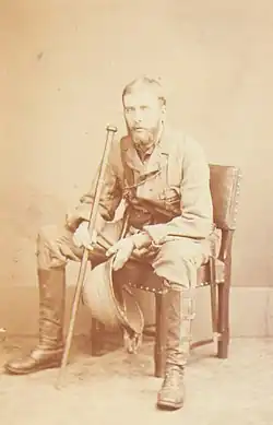 A bearded Russell sits on a chair wearing tropical uniform and holding a cane and a pith helmet
