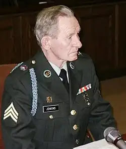 Three-quarters photo of an older white man wearing a formal US Army uniform bearing sergeant's stripes; he is seated behind a table and microphone looking to the right of frame