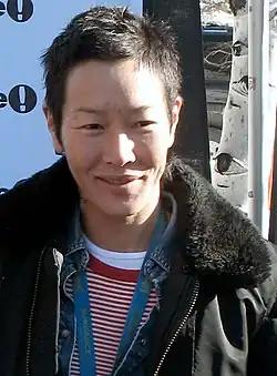 An Asian woman with short dark hair, wearing several layers and a Sundance Film Festival lanyard, smiles with a birch tree in the background