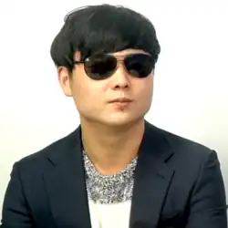 A portrait photograph of young Asian man with short, black hair. He is wearing a pair of aviator sunglasses and a black suit jacket over a white sweater.