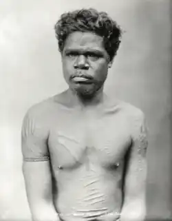 Portrait photograph of a shirtless Aboriginal man with a heavily scarred torso