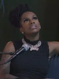 An African-American woman in a sleeveless black top, several necklaces, and earrings sits on a blue couch in front of a microphone and glances upwards