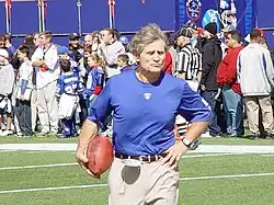 Candid photograph of McNally wearing a blue t-shirt bearing a New York Giants logo standing on a football field and holding a football in his right hand with his left hand on his hip