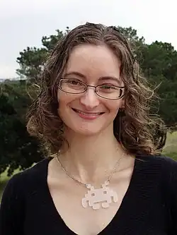 Shoulder-up outdoor portrait of Jo Anderton smiling, wearing glasses, a dark top, and a geometric pendant necklace, with trees blurred in the background.