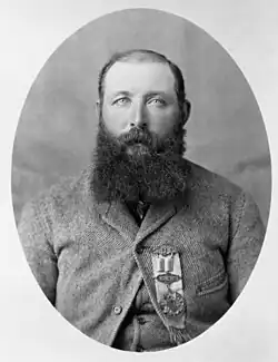 A black and white head and shoulders photograph of a balding white man with a full, dark, beard and a heavy wool coat