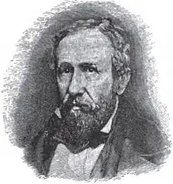 A man with receding black hair, a mustache, and a beard wearing a white shirt and black jacket