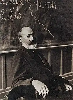 A man in upper middle age, in a professor's studio, wearing glasses. He has a neat beard.