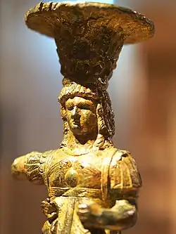 Close-up photograph of a gilded bronze figurine depicting an ancient deity in intricate armor, wearing a vase-shaped headdress. The setting is warm and softly lit.