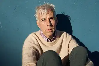 John Vanderslice sitting in front of a teal blue wall