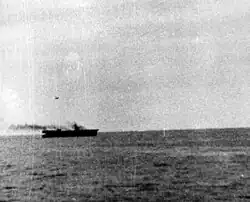 A plane is silhouetted against the sky as it flies downwards towards an aircraft carrier. Smoke running from the funnels shows that the aircraft carrier is making full speed.