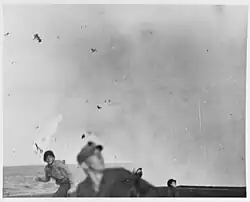 A flaming plane, with its tail pointing towards the ground, is photographed tumbling just above the ocean. In the foreground, standing on top of the flight deck, four men are looking to the right, towards an unseen object. The men have a look of surprise and terror on their faces.