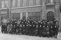 The image shows a large group of people lined up in three rows in the foreground.