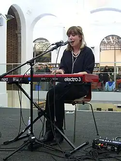 Katie Stelmanis wearing a black outfit with several necklaces, seated, playing a keyboard, appearing to sing into a microphone in what might be a shopping mall, with audience visible in background