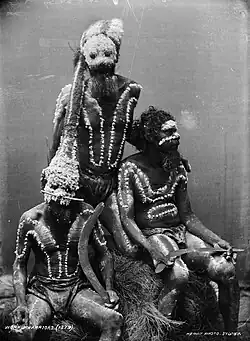 Photograph of three men, two sitting and one standing, wearing headdresses and painted decoration on their torsos. The two men who are sitting are holding boomerangs.