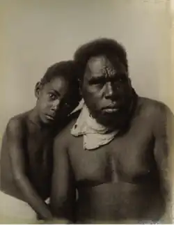 Photograph of an Aboriginal child sitting next to an Aboriginal man, the child's head resting on the man's shoulder