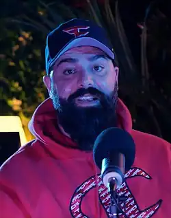 A man with a long beard, black cap, and red hoodie in front of a microphone