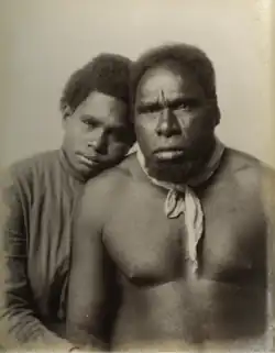 Photograph of an Aboriginal woman sitting beside a shirtless Aboriginal man, her head resting on his shoulder