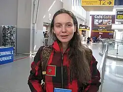 A woman with long brown hair faces the camera, she wears a red and black checked jacket.