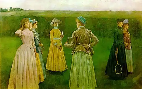 A group of women in long dresses and hats stand in a grassy field holding early tennis rackets, arranged in pairs as if preparing for a match