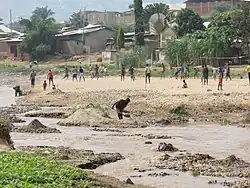Photograph of a group of people playing soccer on the banks of a river next to a village. There is also a man digging sand next to the river in the foreground and a woman washing laundry on one side.