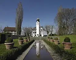 A tall, white, cylindrical tower attached to a chapel-like building is centered within quaint trees and landscaping. The tower and blue skies are mirrored down the length of a long, rectangular reflecting pool lined with hedges and potted plants.