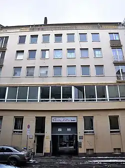 A street-side view of a bland, beige five-story building with rows of windows and occasional balconies. Other than an open garage door, only a single pedestrian door is seen to allow entrance to the building. A sign above the garage says "Elektro-Müller GmbH"