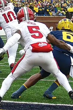 Current New England Patriots cornerback Kobee Minor plays for the Indiana Hoosiers against Michigan in 2023. He is wearing a white Indiana Hoosiers football jersey with his number, 5, and last name, Minor, in red text (last name is smaller and on top of the number). He is also wearing a red helmet with white stripe down the middle as well.