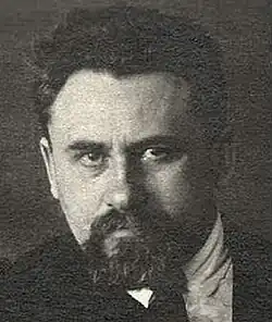 Dramatic, sepia studio publicity portrait of a striking, dark-eyed man looking upward from under heavy brows. He has a trimmed beard, full moustache, and short, curly, dark hair.