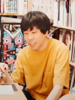 A man with black hair and a yellow shirt, sitting at a table