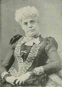 B&W portrait photo of an elderly seated woman wearing a dark, high-collared blouse with her hair in an up-do.