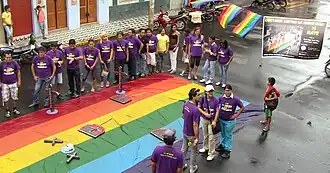 People in purple t-shirts gathered around a large rainbow flag spread on the ground, with additional rainbow flags visible in the background