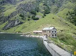 Colour photograph of a concrete dam holding back a lake. In the background, a refuge and mountains.