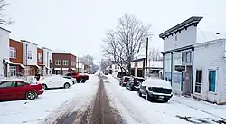 Snow-covered Simpson Street in Lafayette, Colorado, with parked cars lining both sides of the narrow roadway, historic one- and two-story commercial buildings on the right, modern townhouses on the left, bare winter trees, and an overcast sky.