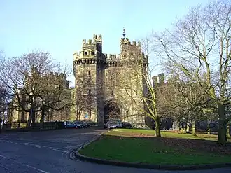 A large stone castle, with imposing towers either side of the gateway, is partly obscured by trees on the green in front of the building. A road leads up to the castle, at the end of which are parked six cars outside the high wooden doors.