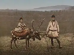 A historic photograph of a Sámi man standing beside a reindeer with a child seated on the animal, taken in Finnmark, northern Norway, around 1890–1900.