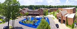 An aerial photograph of the campus featuring a driveway, blue-and-green playground, and several brick buildings. Behind the buildings, only large green trees can be seen.