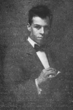 Young man in a dark suit and bow tie, looking at the camera and holding an object in his hands against a dark, grainy background.