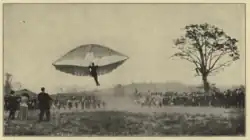 A man hangs below a glider in flight. Many people (at least 100) are watching.