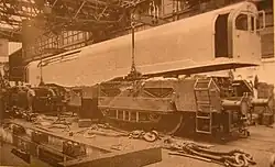 A picture of a locomotive under construction. The cuboid box containing cabs and boiler is being lowered onto the two bogies by a crane inside a locomotive works. Various components are scattered in the foreground.