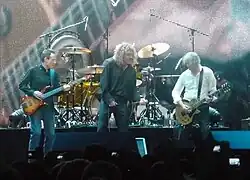 A colour photograph of John Paul Jones, Robert Plant and Jimmy Page performing on stage, with Jason Bonham partially visible on drums in the background