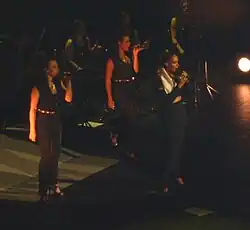 Three women sing on stage. They wear similar outfits.