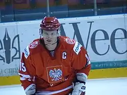 Ice hockey player wearing a red helmet, red and white gloves; and a red jersey with a white Polish Eagle emblem, a captain's letter cee, and the uniform number 15