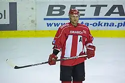 Ice hockey player wearing a red helmet and gloves, black hockey pants; and a red jersey with white stripes and an assistant captain's "A"