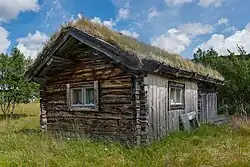 A weathered wooden building with a sod roof and two small windows, set in open grassland with scattered trees.