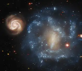 This image shows two galaxies side by side. The galaxy on the top left is smaller in size, and appears as a bright glowing spiral with clearly-defined arms. A larger blue galaxy dominates the full right field of the image. This galaxy is more irregularly shaped, with a glowing central bar, and varying regions of concentrated hues of blue. The background is black with various stars and galaxies in the distance.