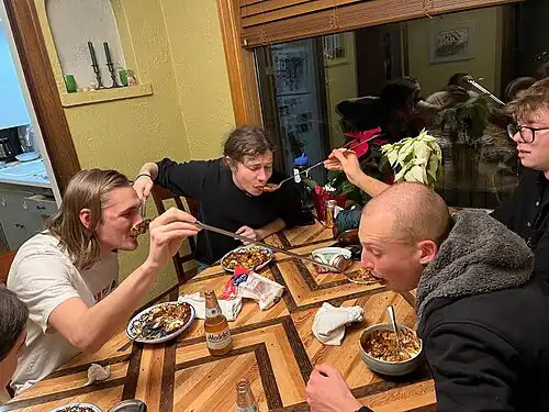 Three young men sat around a dinner table each serve food to the man to their right with a large spoon