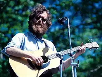 Loudon Wainwright III wearing sunglasses and playing an acoustic guitar on an outdoor stage