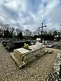 raised rectangular grave with a metal cross at the head. A rectangular urn rests at the base of the cross.